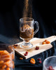 Latvian rye bread dessert in a clear handled glass on a wooden board, with dried fruit and a dusting of spice.